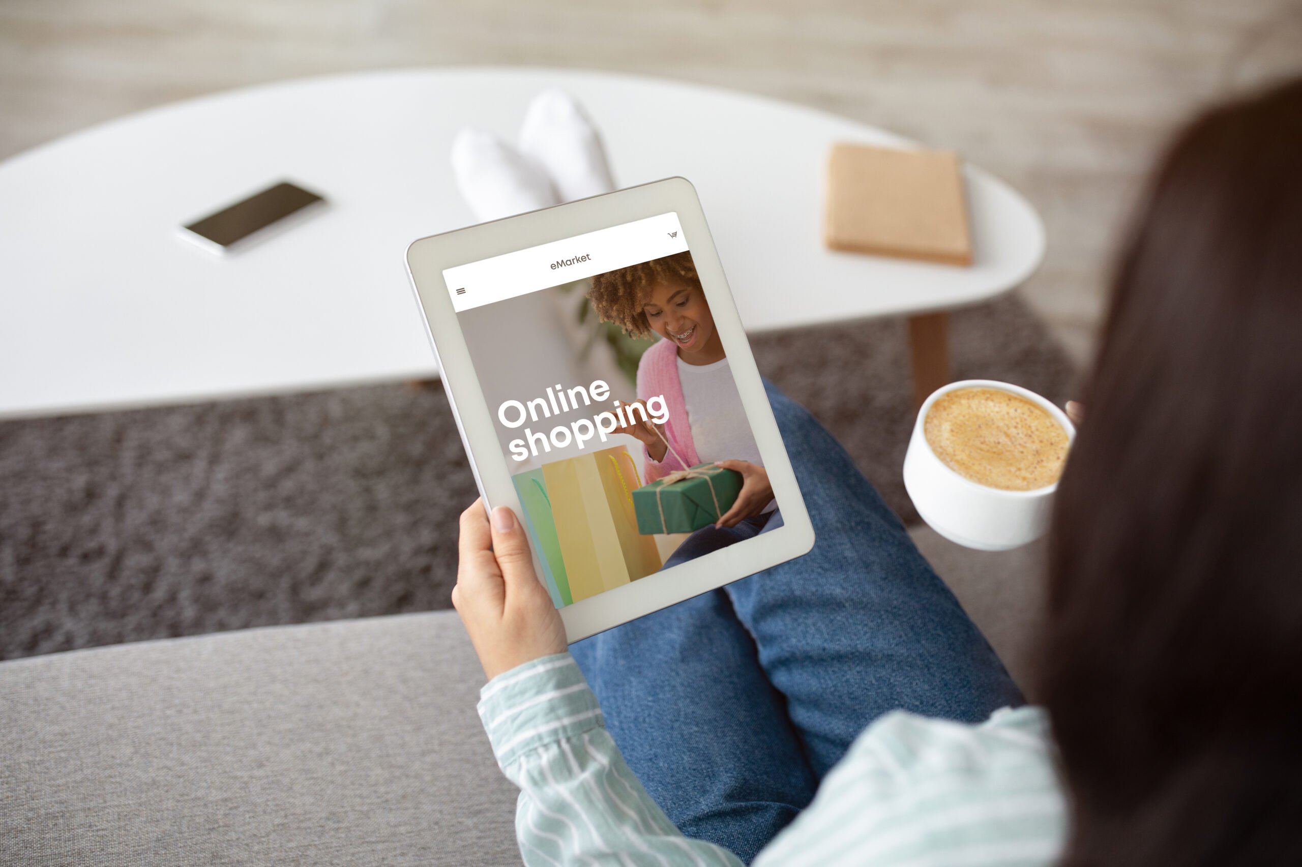 Young indian woman with coffee cup sitting on sofa, using tablet with fashion store on screen at home